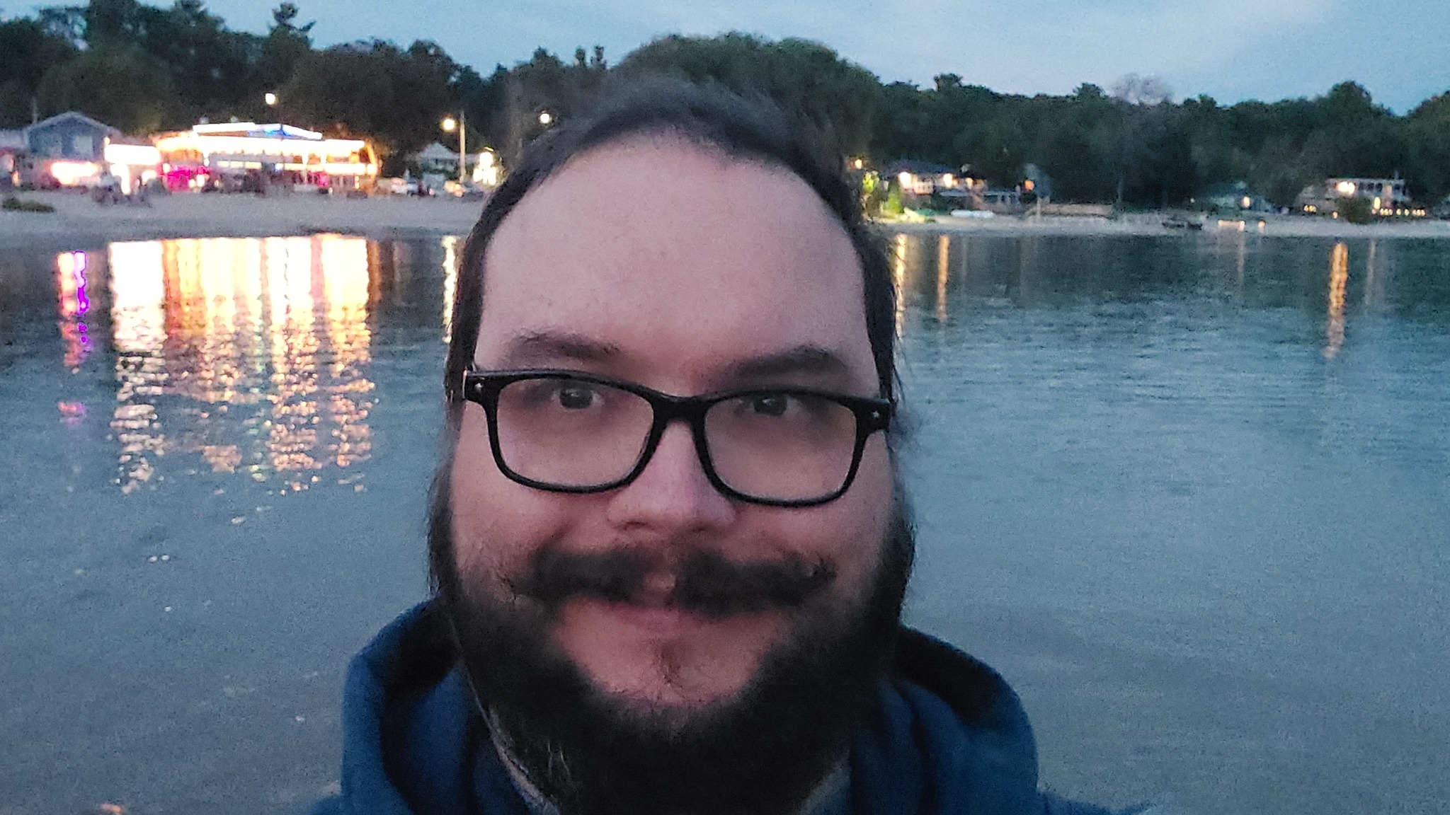 A man with shork dark hair, a beard and moustache stands in front of a bay, brightly lit buildings in the background